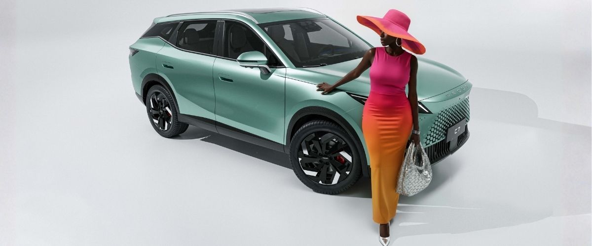 Beyaz stüdyoda çağla yeşili Omoda 7 ve şık şapkalı, turuncu-pembe maksi elbiseli kadın model