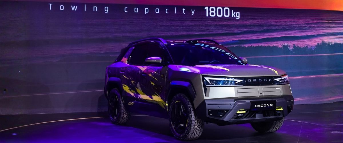 Sahnede sergilenen yeşil ve sarı kamuflaj detaylı yeni OMODA X modeli ve arka ekranda vurgulanan 1800 kg çekme kapasitesi (Towing capacity 1800 kg) özelliği.